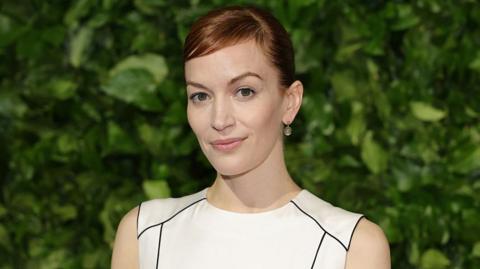 Britt Lower at a red carpet event. She is standing in front of a dense wall of green foliage, Her hair is a vivid shade of red, styled neatly back, She is wearing a sleeveless white dress with black piping that traces the seams and small drop earrings.