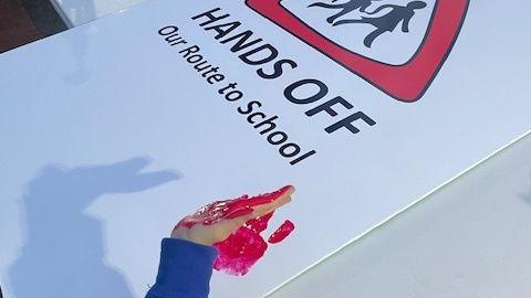 The 'Hands off our route to school' sign before it has been covered in red handprints. A child's arm in a blue jumper can be seen hovering over the sign, with their hand covered in red paint. There is a single red handprint on the sign. 