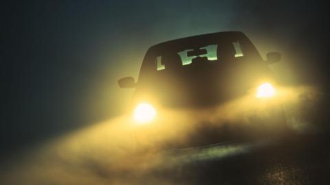 A foggy and dark night with a silhouetted car with headlights casting a glow across the road