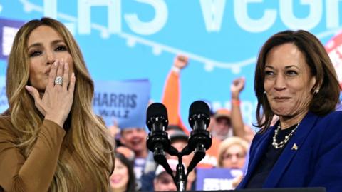 Jennifer Lopez and Kamala Harris on stage. Lopez is blowing a kiss to the crowd.