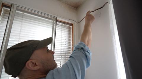 A man in a baseball cap points to a crack in the wall above his window
