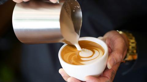 A barista pouring milk foam into an espresso in a white cup, resulting a pattern design on the surface of a latte