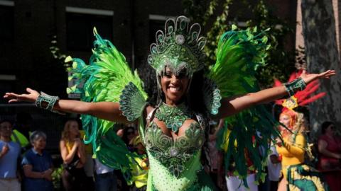 A dancer in green with a flurry of feathers behind her holds her arms out with a wide smile.