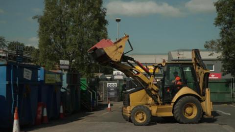 A digger-type vehicle dumping a sofa into a skip at the recycling centre in Totnes. It is being driven by someone in hi-viz.