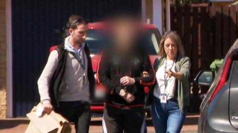 Two plain clothed detectives lead away a man into an unmarked police car in a suburban housing estate. The officers - one male, one female are in jeans and trainers. The suspect is in trainers and tracksuit bottoms and has been blurred to hide his identity.