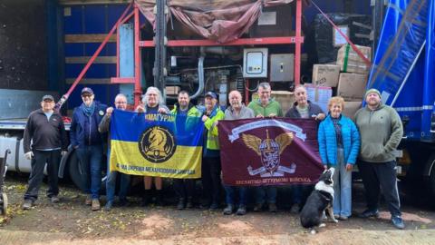 Volunteers of Macclesfield Ukrainian Aid hold banners as they stand in front of a lorry loaded with aid destined for the country