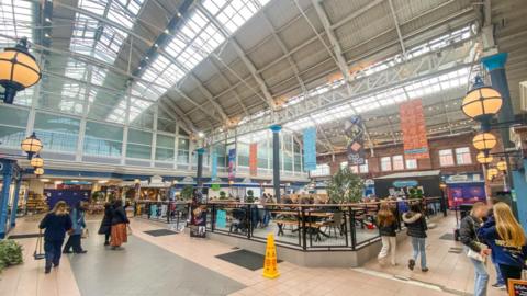 A general view of the refurbished food court at Carlisle Market Hall and Eden Centre. The large space has a vaulted ceiling with skylights. Stalls are placed all around a central area with picnic style wooden benches, busy with people eating and drinking. There are colourful banners hanging from the ceiling and large round lantern-type lights dotted around the space.