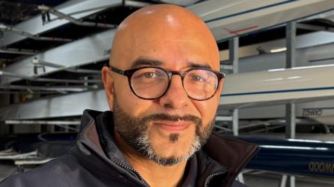 Omar Terywall, centre frame and looking directly out of the image. He is smiling slightly, has a dark, neatly trimmed beard, flecked with grey on his chin, and is wearing circular, black-framed glasses. He is bald. He is wearing a dark blue, collared top, but only the top of one shoulder on the left can be seen. Behind Omar are several sporting rowing boats mounted on racks inside a boathouse.