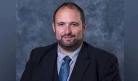 A man with brown hair and a beard pictured in a black suit blazer, blue shirt and navy blue tie. He is slightly smiling and against a blue backdrop.