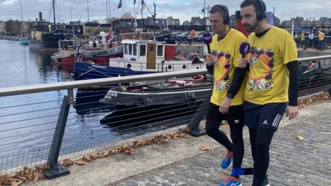 Ben Watts and Adam Dowling wearing Children in Need t shirts, carrying microphones and wearing headset walking three-legged around St Mary's Island with boats and water in the background
