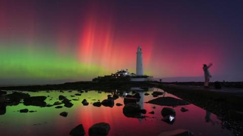 The aurora borealis, also known as the northern lights, glow in the sky over St Mary's Lighthouse in Whitley Bay on the North East coast, reaching a G4 level geomagnetic storm, putting on an amazing display in the early hours. The lights are pink and green and reflect off the water, a woman stands next to the water taking a picture with the white lighthouse in the background.