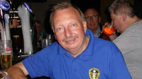 Michael leans on a bar in a dark pub. He wears a blue Leeds United branded polo shirt. He has short hair and a moustache.