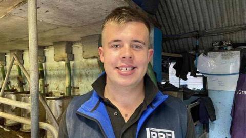 Jack Emery smiling at the camera has blonde hair and blue eyes. He is wearing a black T-shirt and a blue fleece. In the background is a cow milking parlour.