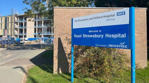 A blue and white sign outside a hospital site that reads "welcome to royal shrewsbury hospital. this hospital is smoke free". behind it is a small brick building. in the background is the hospital building with lots of scaffolding and a white crane above it.