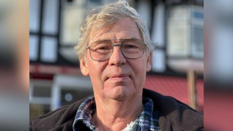 A close-up head and shoulders picture of a man wearing rectangular thin grey glasses. He has short grey hair and is wearing a brown jacket and a blue and green checked shirt