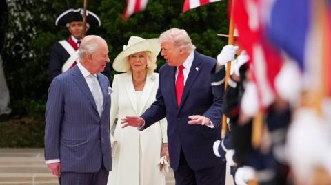 The King, Queen and Trump walk outside the White House with UK and US flags in the foreground.