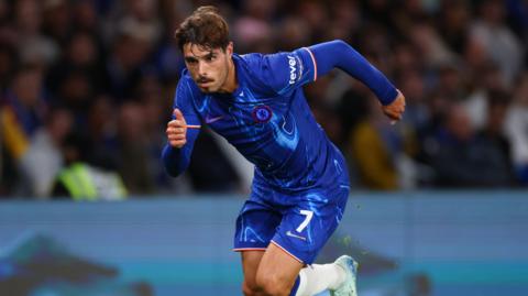 Pedro Neto, wearing Chelsea's home kit of all blue with a white 7 on his blue shorts runs for the ball, which is not in shot, with a blurred background of fans behind him