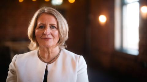 Anne Longfield has bobbed blonde hair. She is wearing a formal white jacket.