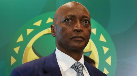 A headshot of Patrice Motsepe wearing tie and suit standing in front of a Confederation of African Football banner