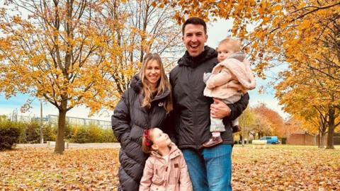 Lucy and Tom Fitton with their two daughters. The family is standing in a yellow and orange leaf strewn grassy area, with trees on either side. They are all smiling.  Lucy is wearing a long black padded coat, Tom, who is holding his youngest daughter, is wearing blue jeans and a black waterproof jacket and the girls are wearing pale pink coats. 