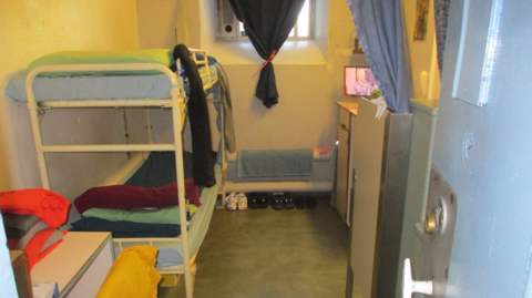 A cramped cell with bunk bed and shoes on the floor. There are small cupboards to the side and objects on top of cupboards.