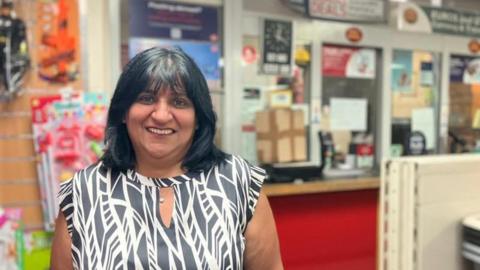 A woman with black shoulder-length hair wearing a black and white top smiles at the camera. She stands in a office near the counters.