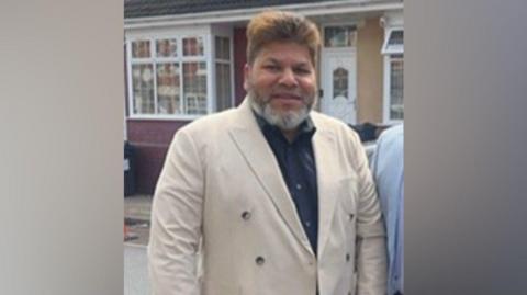 A man with brown hair and wearing a white jacket over a black shirt, standing on a street in front of a house. He has a grey and black beard.