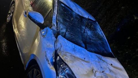 The windscreen of a white car is smashed and the bonnet is bent. 