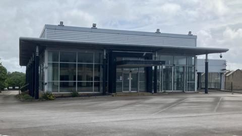 Former Mercedes car dealership building with glass doors and a concrete yard outside.