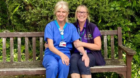 Two women sitting close together on a bench outside, smiling at the camera. One of them is wearing blue hospital scrubs. The other one is wearing a purple short-sleeved top and dark trousers.