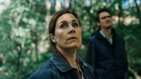 A woman looks up apprehensively in the foreground, while a man looks up in the background, with green foliage visible behind them both
