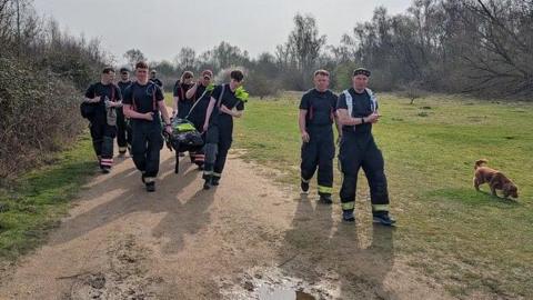 A group of nine young people, wearing dark coloured t-shirts and trousers with fluorescent stripes towards the bottom walking along a path towards the camera. Some of them are holding up a stretcher with a dummy lying on it. Around them is grass and trees. There is also a small dog on the grass. 