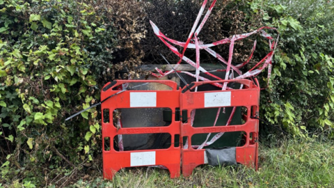 A communications box blackened as the result of a fire, with red safety barriers in front of it and red and white fire tape around it. The box is in front of several green bushes.