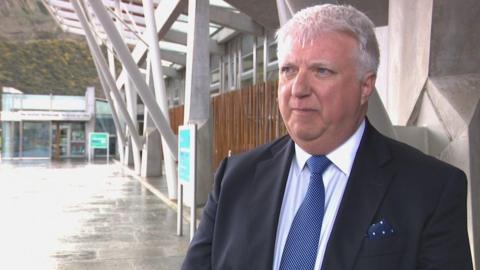 David Harris, a man with white hair, wearing a dark suit and a blue tie, standing outside the Scottish parliament building in Edinburgh