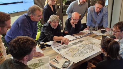 People sat around a blueprint of a building on a table, pointing and listening to each other