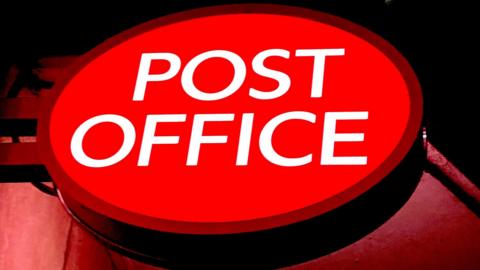 An illuminated red, oval shaped sign with the words "Post Office" in white capital letters.