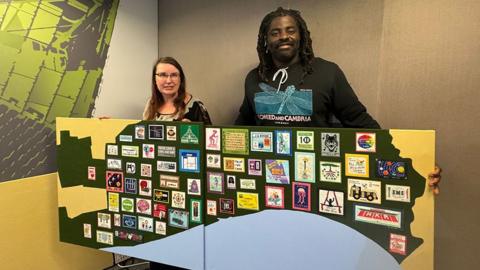 Miranda van Rossum and Kofi Smiles holding the embroidery it has a black outline of Hull next to the blue of the Humber Estuary with brightly coloured stiches squares arranged across it.