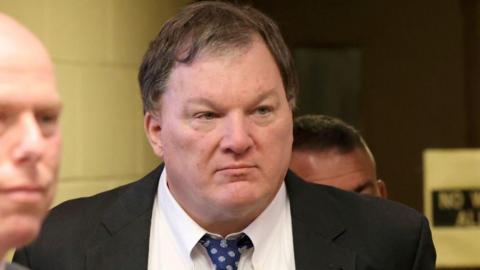 A close photo of Rex Heuermann, 62, in a courtroom. He wears a black jacket and blue patterned tie