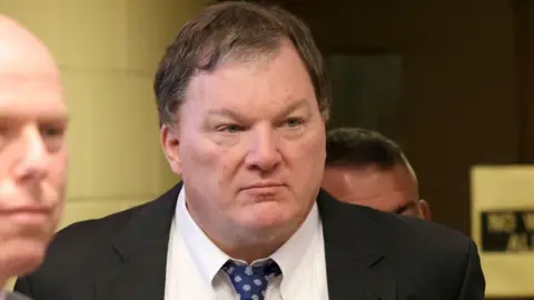 A close photo of Rex Heuermann, 62, in a courtroom. He wears a black jacket and blue patterned tie