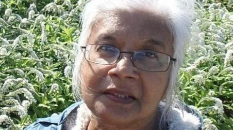 A family handout of Mena. She has long white hair and is wearing glasses. She is standing in front of a garden.