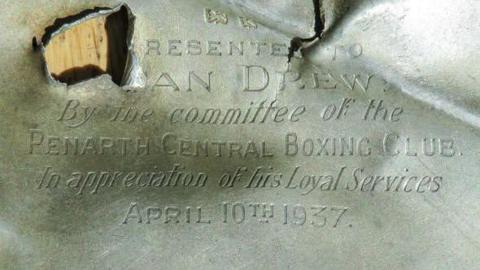 A buckled silver cigarette case: the top right is folded in upon itself, whilst in the top left a bullet hole obscures the inscription