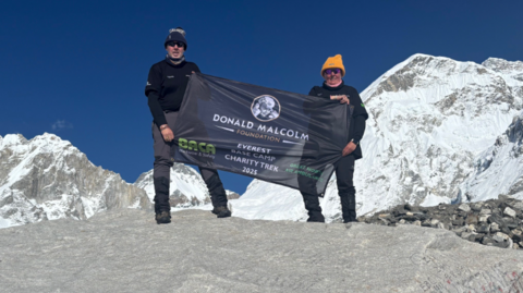 The photo shows two people standing on a large boulder in a high‑altitude mountain landscape, surrounded by snow‑covered peaks under a blue sky. They are holding a black banner that reads “Donald Malcolm Foundation – Everest Charity Trek 2025” along with additional text and logos.