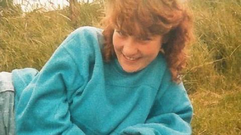 A girl with brown, curly hair is wearing a blue jumper and is seen smiling whilst laying on her side in long grass.