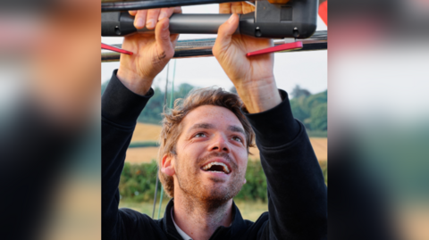 Hugo Hall holding on to a balloon burner. He is looking up and has an excited look on his face