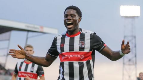 Grimsby Town winger Justin Amaluzor holds arms out wide in celebration with wide smile wearing home black and white kit