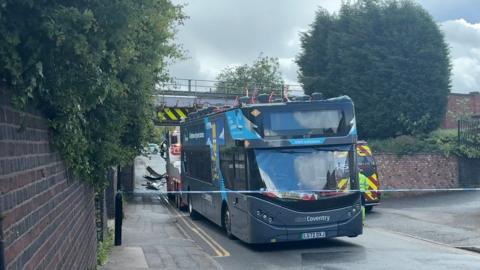 A grey double decker bus with its roof torn off is stationary in the road. Behind it the bridge is the railway bridge and debris left on the road.