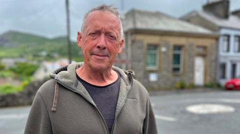 Gruffydd looks at the camera. He has short grey hair, and wears a khaki zip fleece with a black top underneath. A mini roundabout and a stone building can be seen in the background.