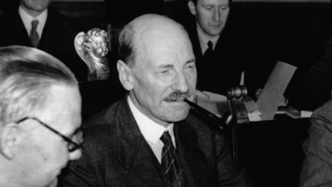 A Black and white photo of prime minister Clement Attlee looking serious while smoking a cigar and wearing a suit. He is holding a box of matches in his right hand above the table. He is sat in a meeting behind a table surrounded by other men at the Potsdam Conference in 1945.