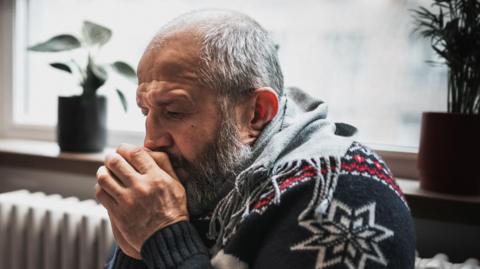 A man in a Christmas jumper huddles for warmth next to a heater