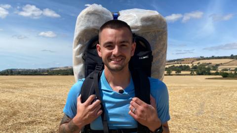 Photograph of Firmstone, who is looking into the camera and smiling. He is wearing a bright blue T-shirt, and has a hay bale wrapped in plastic sheeting attached to a backpack on his back. He is holding onto the chest straps, and is stood on a yellow, stubbly field. The sky is bright blue and there is strong sunshine. 